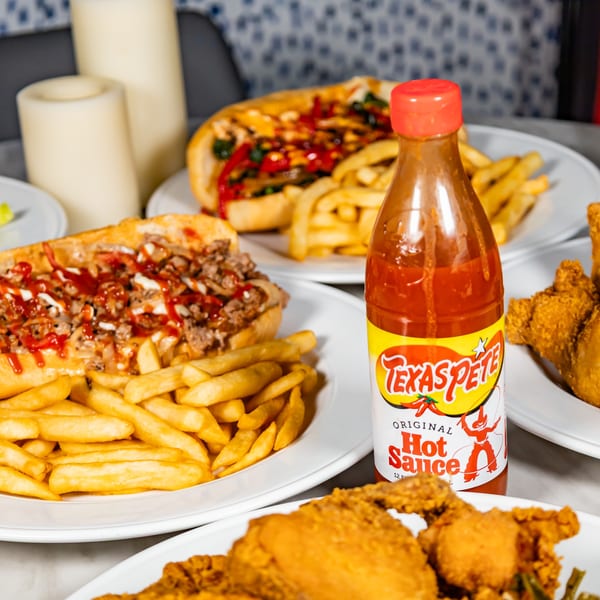 Cheesesteak sandwiches, french fries, fried chicken, and a bottle of Texas Pete hot sauce on a table.