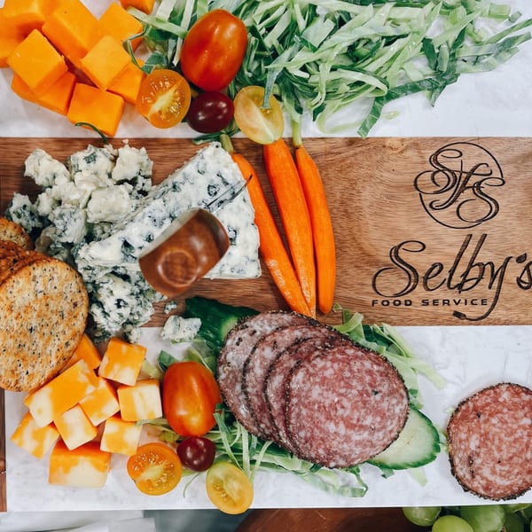 Selby's charcuterie board with cheeses, crackers, salami, carrots, tomatoes, and greens.